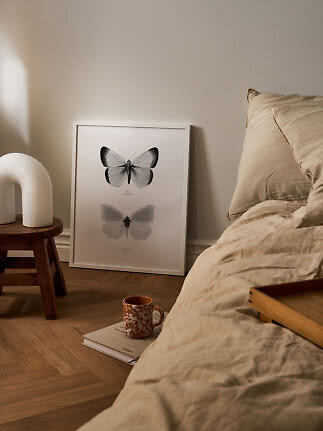 Indrammet sommerfugleplakat lænet mod væg ved seng, med skammel, lampe og krus på gulvet