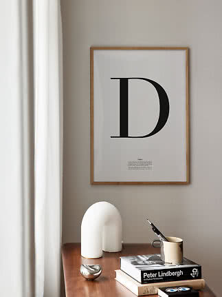 Poster tipografico incorniciato con la lettera D sopra un tavolo in legno con libri e tazza