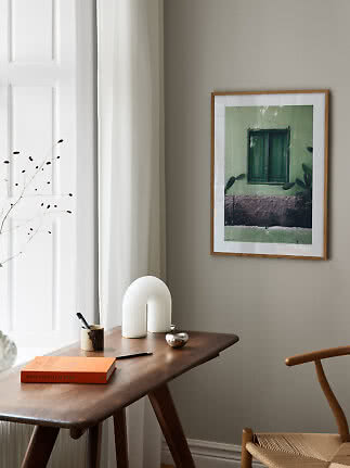 Ingelijste poster met groen raam boven houten bureau met lamp, boek en stoel bij gordijn