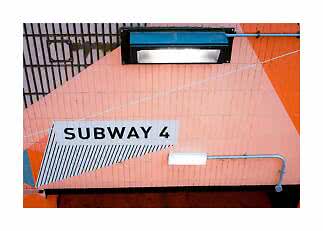 Schild mit „SUBWAY 4“ an einer rosa-orangen Fliesenwand mit Lampen und sichtbaren Rohren