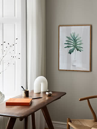 Indrammet plakat med grøn monstera-blad over et træskrivebord i et lyst, minimalistisk rum