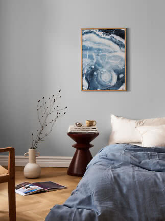 Schlafzimmer mit blauer Bettwäsche, dunklem Beistelltisch, Vase mit Zweigen und abstraktem blauem Wandbild