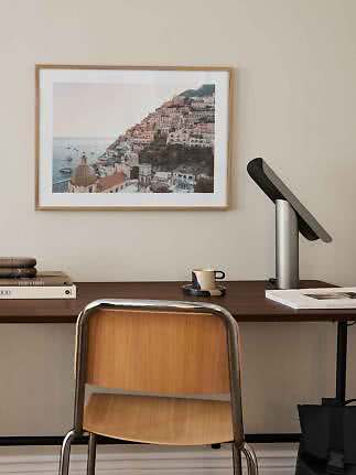 Framed coastal hillside town print above a desk with lamp, cup, and chair