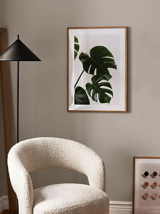 Lámina botánica enmarcada con hojas verdes de monstera en pared beige sobre una silla color crema.