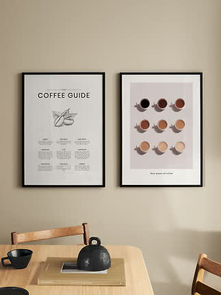 Two framed coffee posters above a wooden table with a black cup, book, and black vase.
