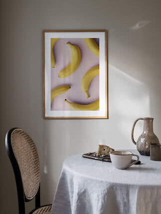 Indrammet plakat med gule bananer på lyserød baggrund over et dækket bord