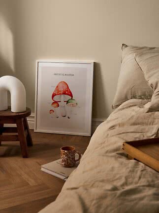 Framed mushroom poster leaning by bed, with white lamp on stool and mug on a book