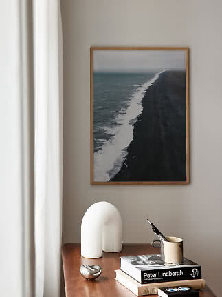 Indrammet kystfotografi over sort sandstrand på beige væg over et træbord med bøger og dekoration