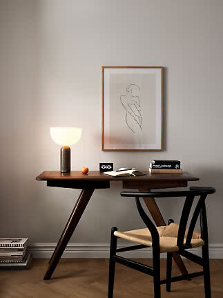 Dark wood desk with glowing table lamp, books, open notebook and black chair beneath framed line art.