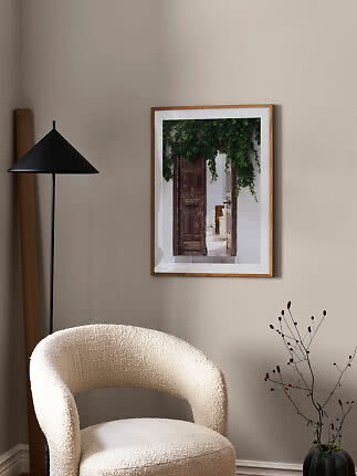 Póster enmarcado de una puerta rústica con vegetación, en pared beige sobre sillón crema