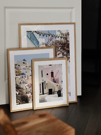 Three framed travel prints in light wood frames leaning against a white wall on a dark floor.