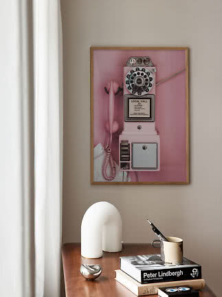 Lámina enmarcada de un teléfono público vintage rosa sobre una mesa de madera con libros y decoración