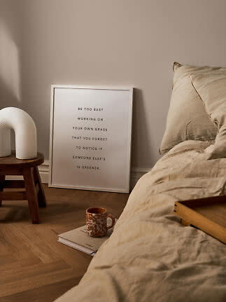 Minimalistisk soveværelse med beige sengelinned, indrammet citatplakat, hvid lampe på skammel og krus på bog