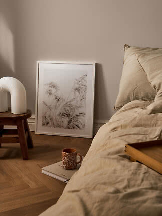 Gemütliches Schlafzimmer mit beiger Bettwäsche, weißer Lampe auf Hocker und gerahmtem Gräserbild