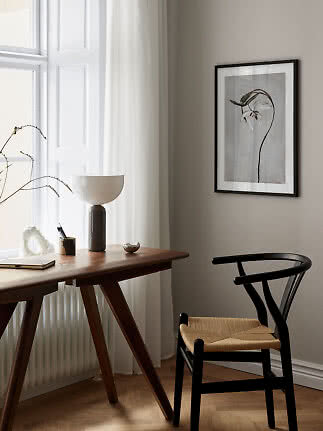 Minimalistisk hjemmekontor med træbord, sort stol, bordlampe og botanisk plakat ved vindue.