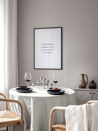 Minimalistisk spiseplads med rundt bord, sorte tallerkener, to vinglas og citatplakat på væggen.