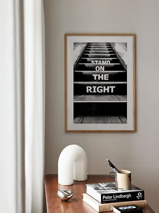 Oprawiony czarno-biały plakat ze schodami ruchomymi i napisem „STAND ON THE RIGHT” na beżowej ścianie