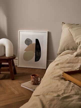 Hyggeligt soveværelse med beige sengetøj, abstrakt plakat, hvid lampe på skammel og krus på gulvet