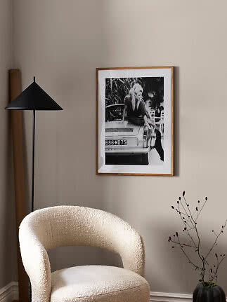 Wood-framed black-and-white poster on a beige wall above a cream chair beside a black floor lamp