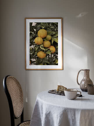 Ingelijste poster met sinaasappels aan een tak boven een gedekte tafel.