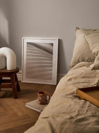 Cozy bedroom with beige bedding, wooden stool, white lamp, and framed print on the floor