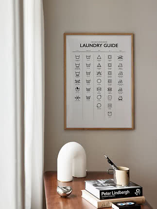 Gerahmtes Wäscheguide-Poster über einem Holztisch mit Büchern, Tasse und weißer Deko