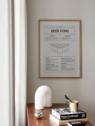 Indrammet Beer Pong-plakat over træbord med bøger, vase og krus ved et vindue