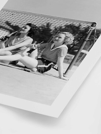 Black-and-white photo of two women by a tennis net holding soft drinks