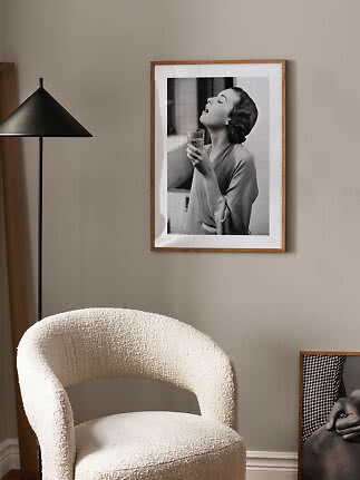 Framed black-and-white poster of a woman holding a glass on a beige wall above a cream chair