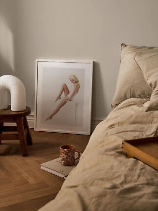 Cozy bedroom with beige bedding, framed art print, white lamp and mug on the floor