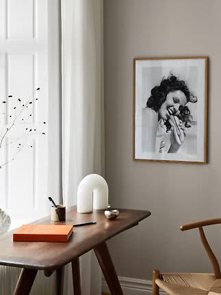 Træskrivebord ved vindue med hvid lampe og indrammet sort-hvidt foto på væggen