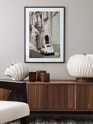 Framed print of a white vintage car on a narrow old street above a wooden sideboard