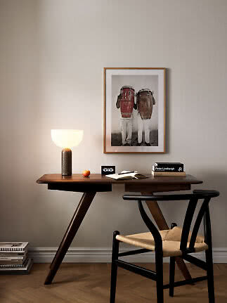 Scrivania in legno scuro con lampada, libri, taccuino aperto e sedia nera sotto quadro incorniciato