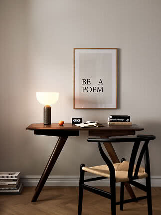 Træskrivebord med tændt bordlampe, stol og indrammet plakat på beige væg