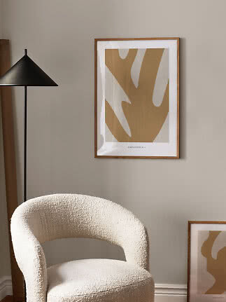 Abstract beige poster in a light wood frame beside a black floor lamp and a white armchair