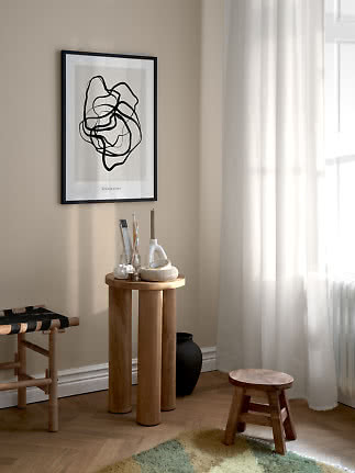 Framed abstract black-and-white poster on a beige wall above a round wooden table by a bright window