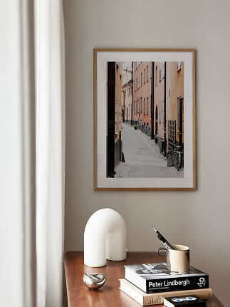 Framed photo of a narrow cobblestone street above a wooden table with books, vase and mug