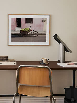 Skrivebord med stol og lampe under indrammet fotokunst af en cykelvogn med bananer