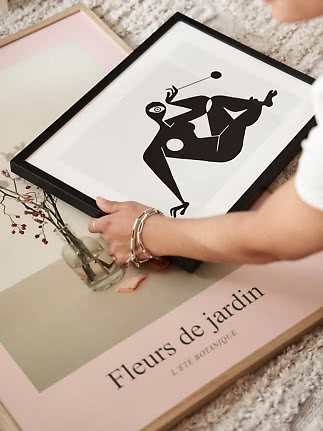 Person hält gerahmten abstrakten Schwarz-Weiß-Druck über rosa botanischem Poster und kleiner Vase