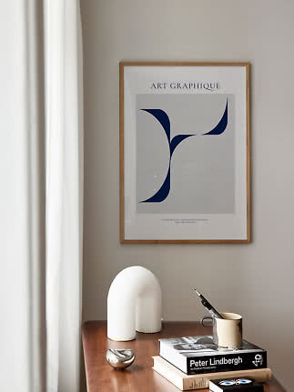 Framed abstract art print above a wooden table with books, mug, white sculpture and metal object