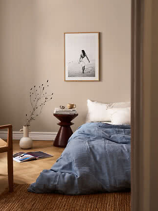 Gemütliches Schlafzimmer mit beiger Wand, Bett mit blauer Decke, Beistelltisch und gerahmtem Strandfoto.