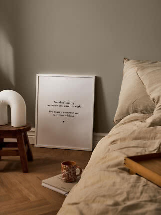 Soveværelse med beige sengetøj, indrammet citatplakat, træskammel med hvid lampe og krus på bog.