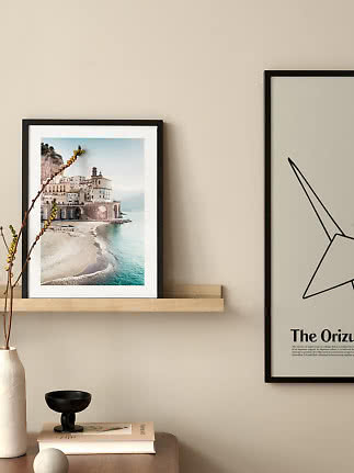 Black-framed coastal print on a wooden shelf above a table with vase, book and black bowl
