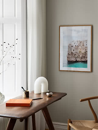 Framed coastal print above a wooden desk with book, pens and white lamp by the window