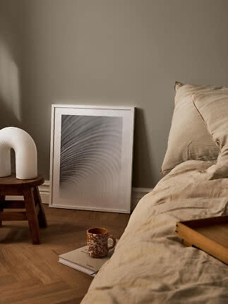 Cozy bedroom with beige bedding, framed art, table lamp and mug on the floor