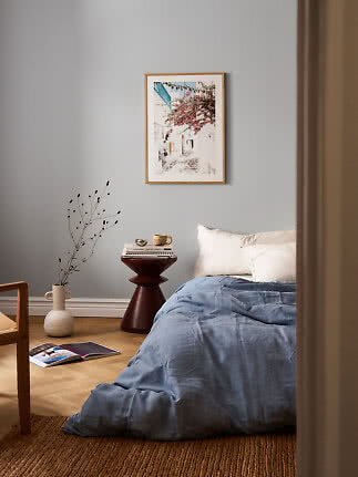 Bedroom with blue bedding, white pillows, side table, vase and framed print on a gray wall