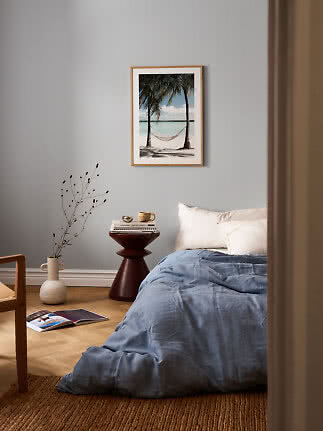 Schlafzimmer mit blauer Bettwäsche und gerahmtem Strandbild mit Hängematte an grauer Wand