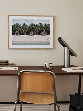 Desk with chair and lamp beneath framed beach photo with palm trees and small huts