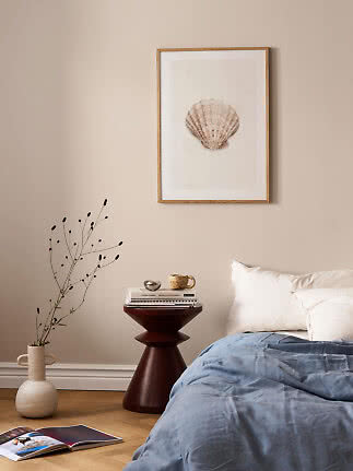 Framed seashell print above a bed in a calm bedroom with side table, vase, and blue duvet