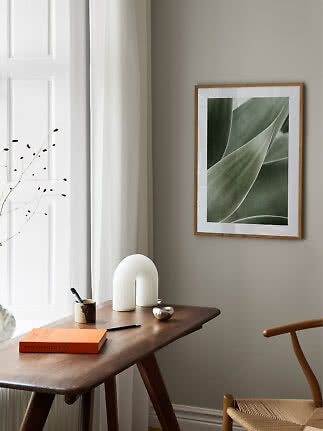 Ingelijste botanische print met groene bladeren boven een houten tafel in een lichte, minimalistische kamer